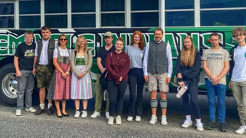 Gruppe aus Lehrern und Schülern - teilweise in Dirndl und Lederhose - steht in den USA vor einem grünen Schulbus