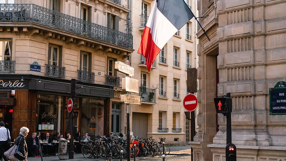 Straße in Frankreich mit hohen Häusern rechts und links, vielen Fahrrädern und Menschen, die im Café sitzen, französische Fahne auf der rechten Häuserseite