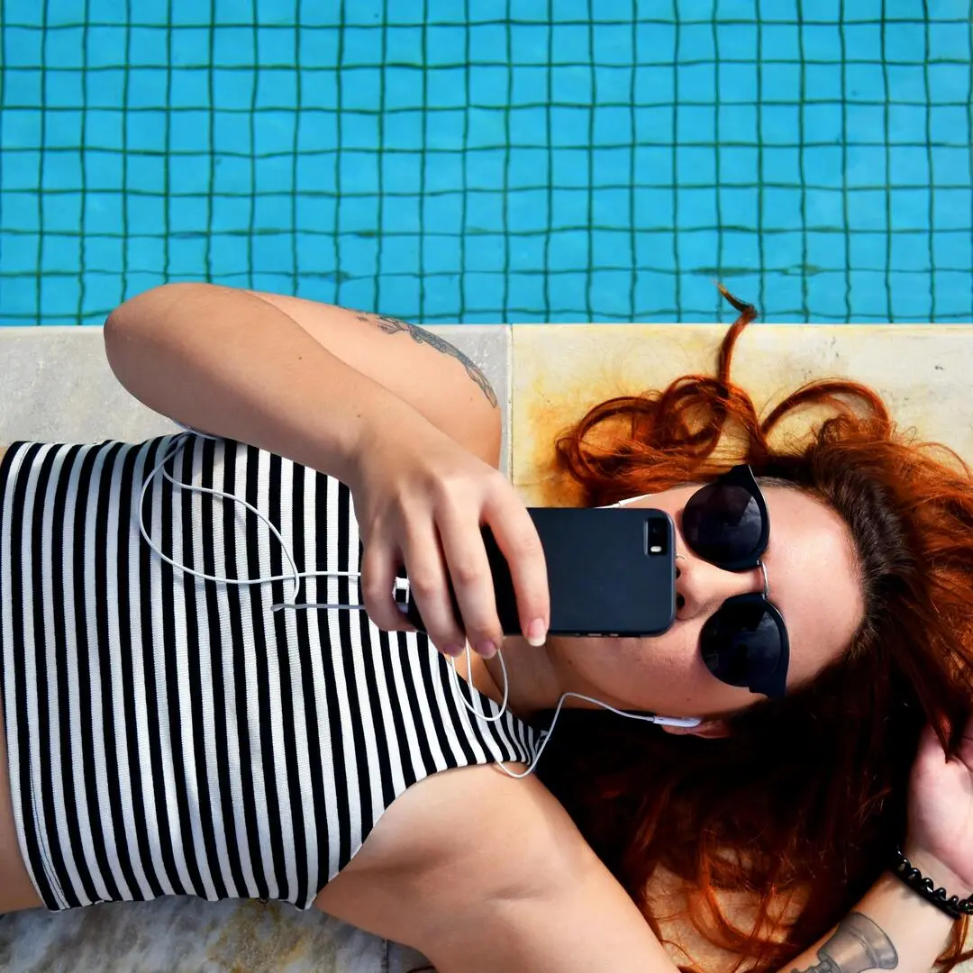 Frau liegt auf dem Boden vor einem Pool und checkt ihr Mobiltelefon